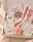 Gift box with pink cozy socks, “Sending Hugs” card, “Proud of You” keepsake, prosecco gummies, star-shaped iced cookie, and self-heating eye mask, styled with soft pastel decor and flowers.