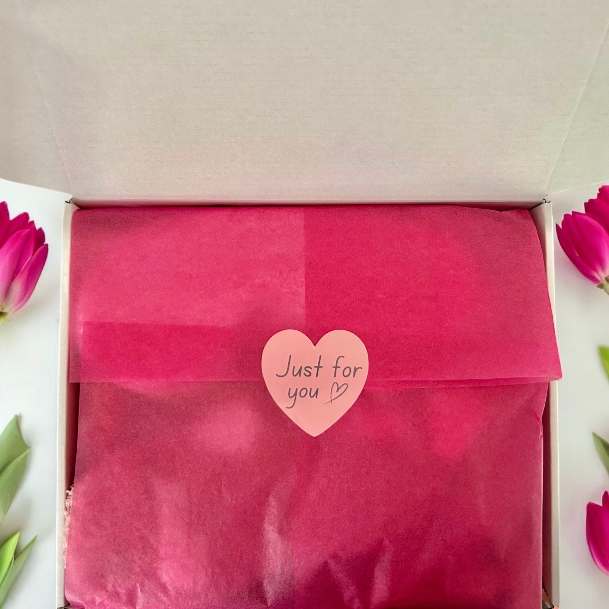 Pink gift bag with 'Just for you' heart-shaped tag in an open box, surrounded by pink flowers.