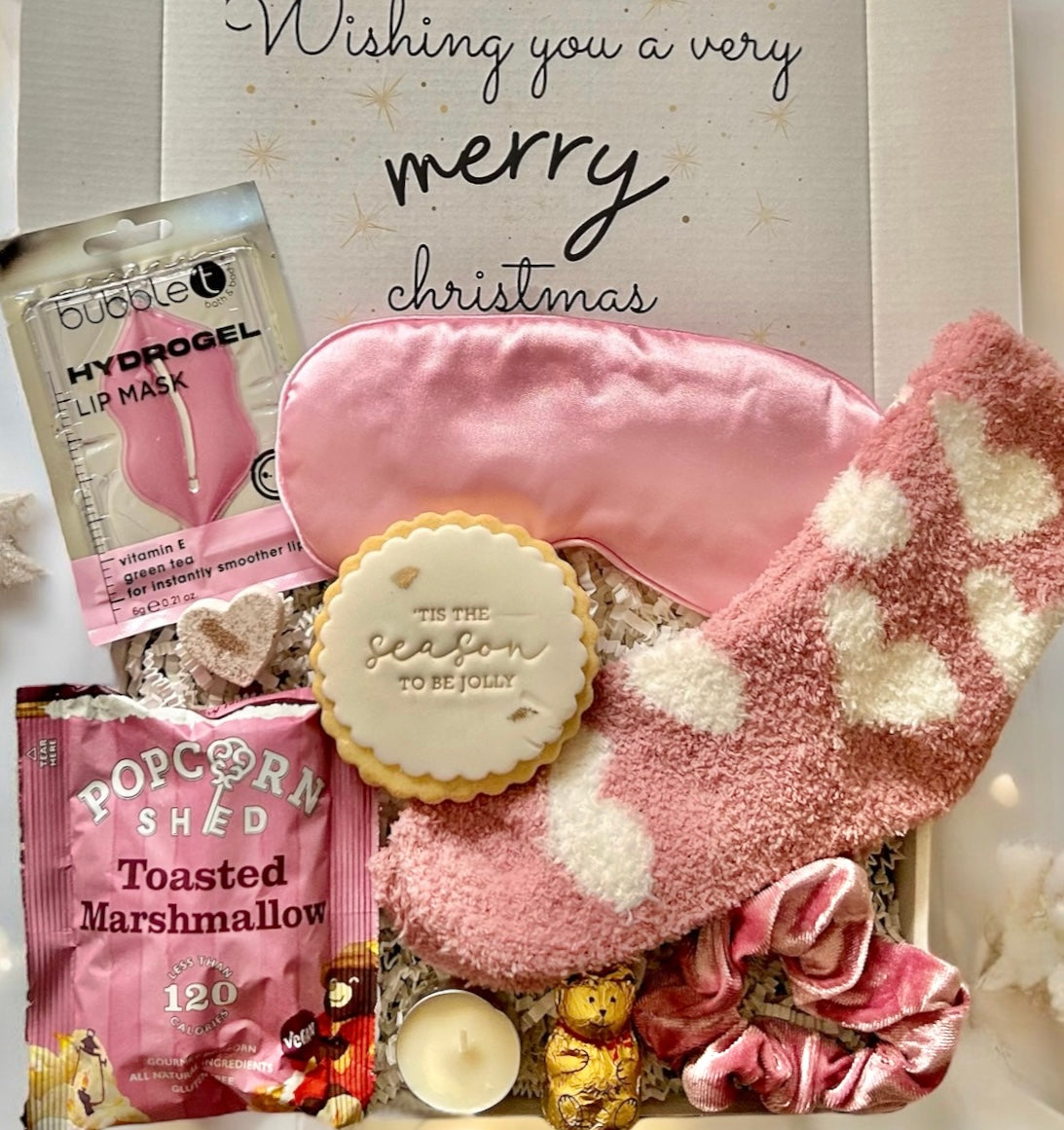 Gift set with pink lip mask, eye mask, popcorn, and cookie with 'Wishing you a very merry Christmas' text.