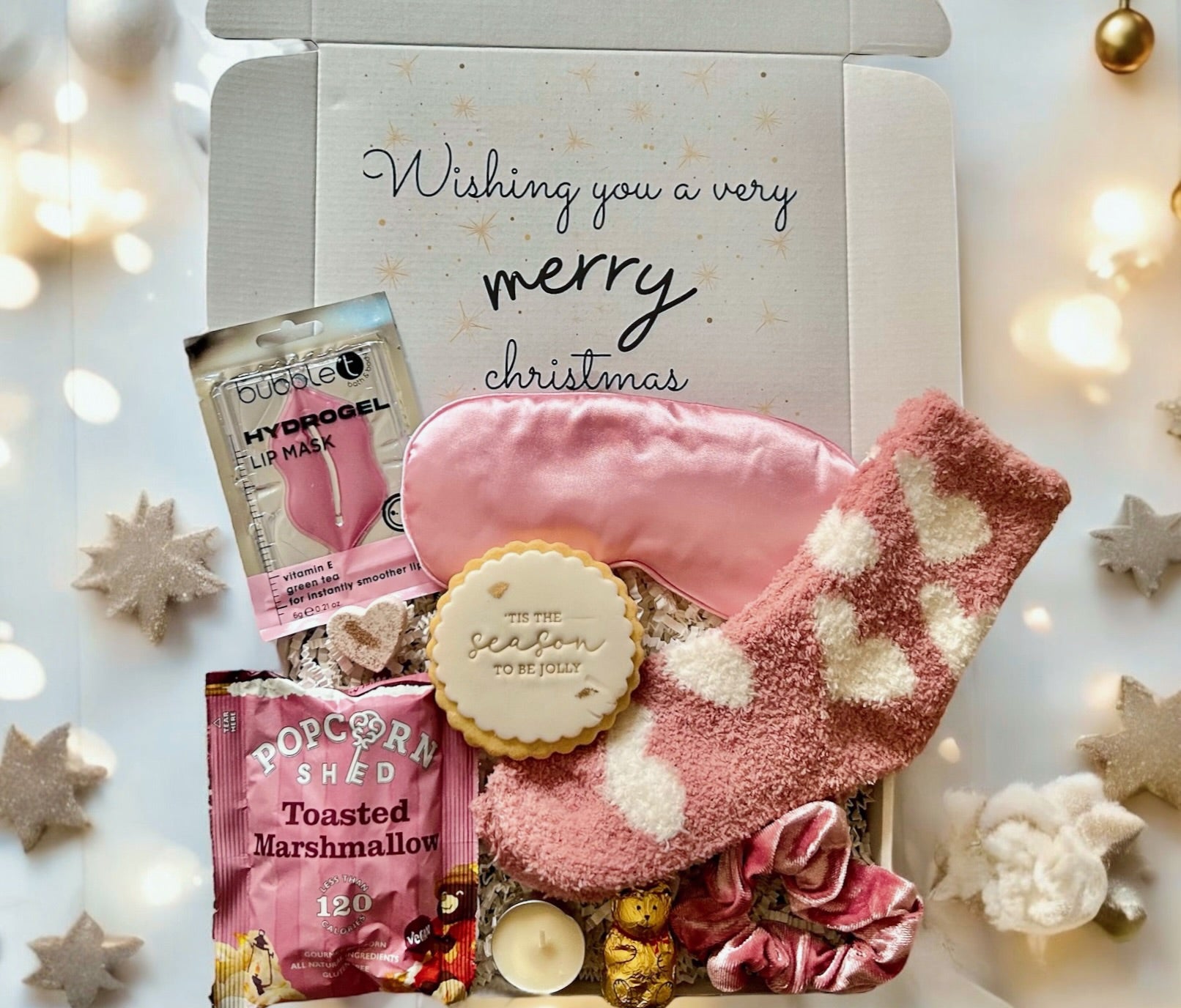 Christmas gift box with pink socks, cookies, and snacks on a white background with festive lights.